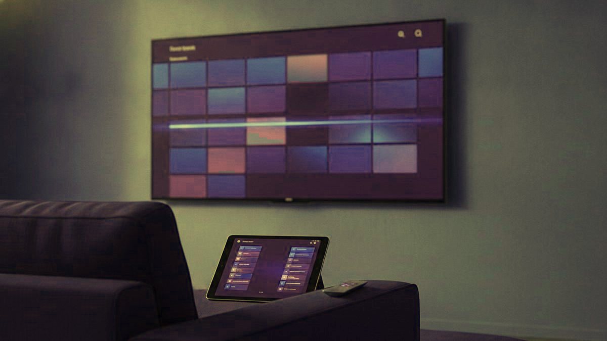 A TV showing a program guide grid with a tablet and phone showing channel lists on a sofa