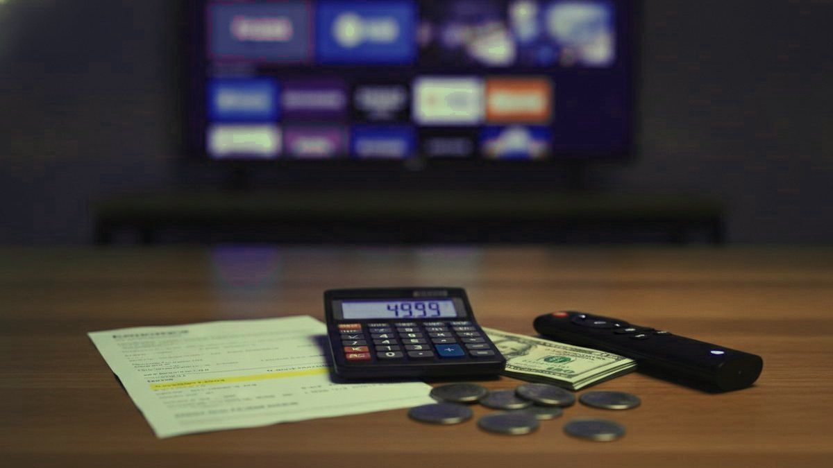A calculator, dollar bills, coins, a streaming stick and a remote on a table in front of a TV